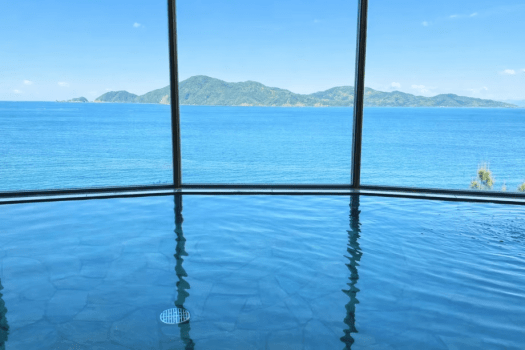 【悠久の湯／オーシャンビュー展望大浴場】長門五名湯に数えられる美肌の湯。そのやわらかな湯面の先に広がるのは、青海島と深川湾の穏やかな海景。ここでしか味わえない、静かな安らぎが満ちていく。