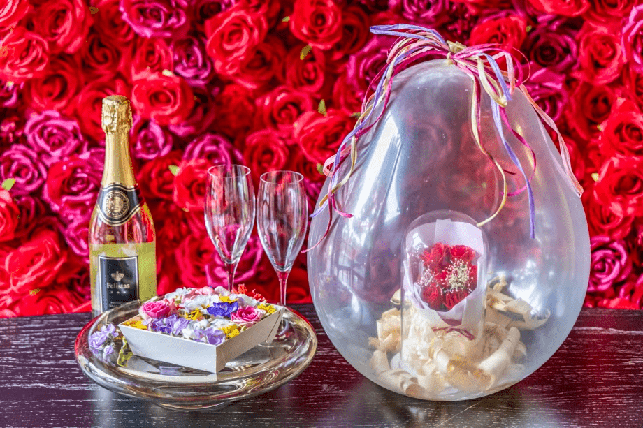 【アニバーサリープラン】華やかなローズウォールを背景に、シャンパンとグラスをご用意。さらに、バルーンの中に包まれた花束と彩り豊かなフラワーボックスが、特別な一日を優雅に演出